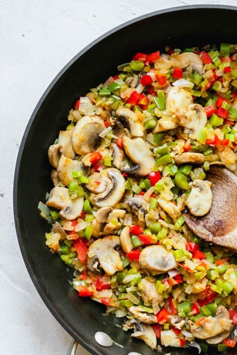 mushrooms, celery, onions, and bell peppers in a large skillet with a wooden spoon