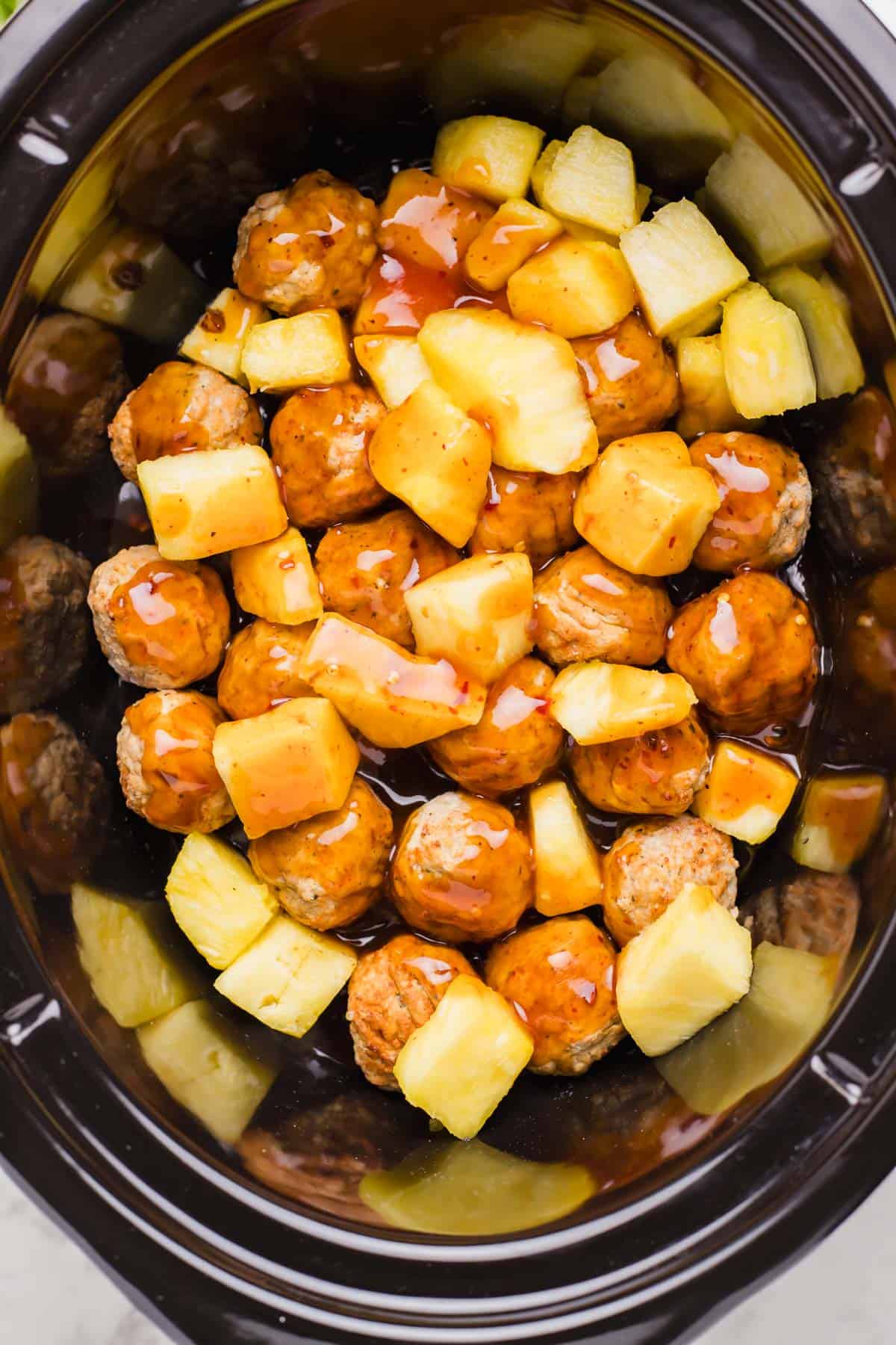 sweet and sour sauce poured over pineapple and meatball mixture in the insert of a slow cooker