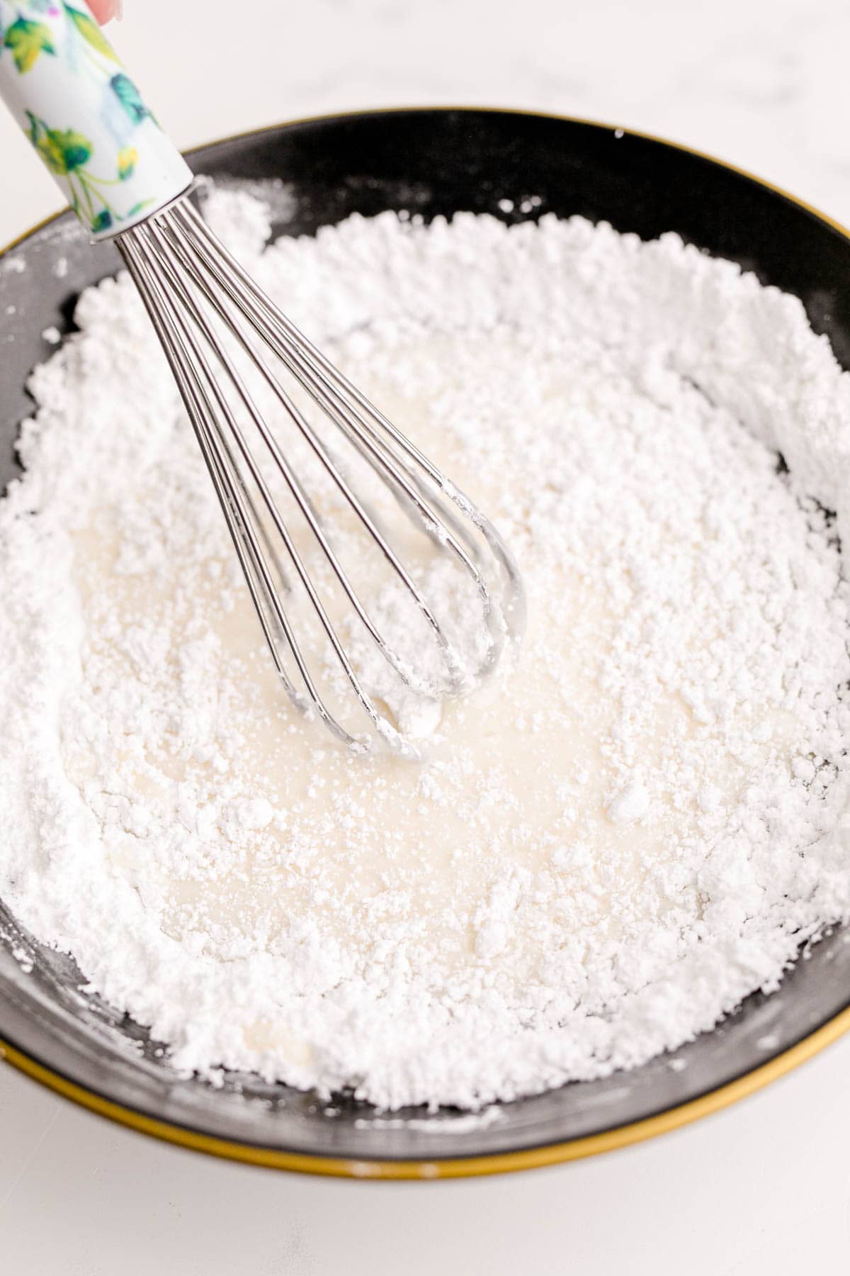 powdered sugar in a black bowl with liquid and a metal whisk