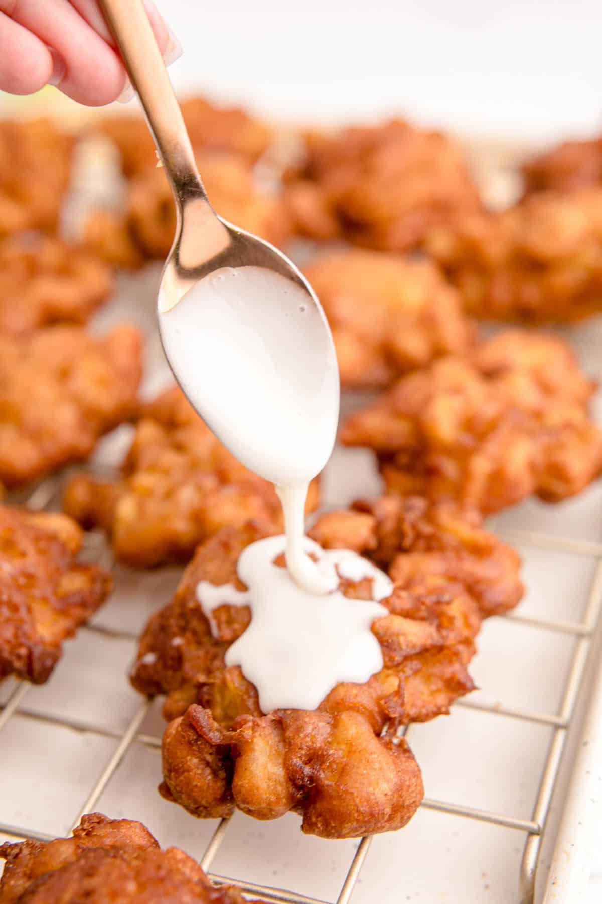 drizzling glaze from a gold spoon on top of fried apple fritters on a wire rack