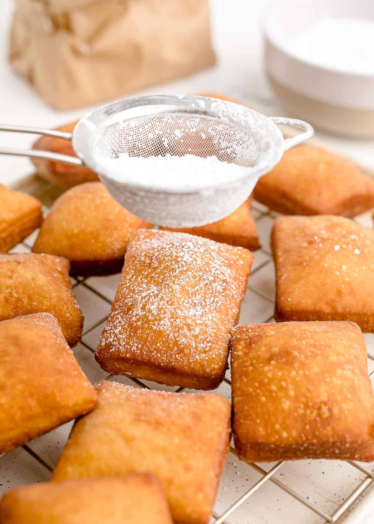 dusting powdered sugar on fried beignets cooling on a wire rack