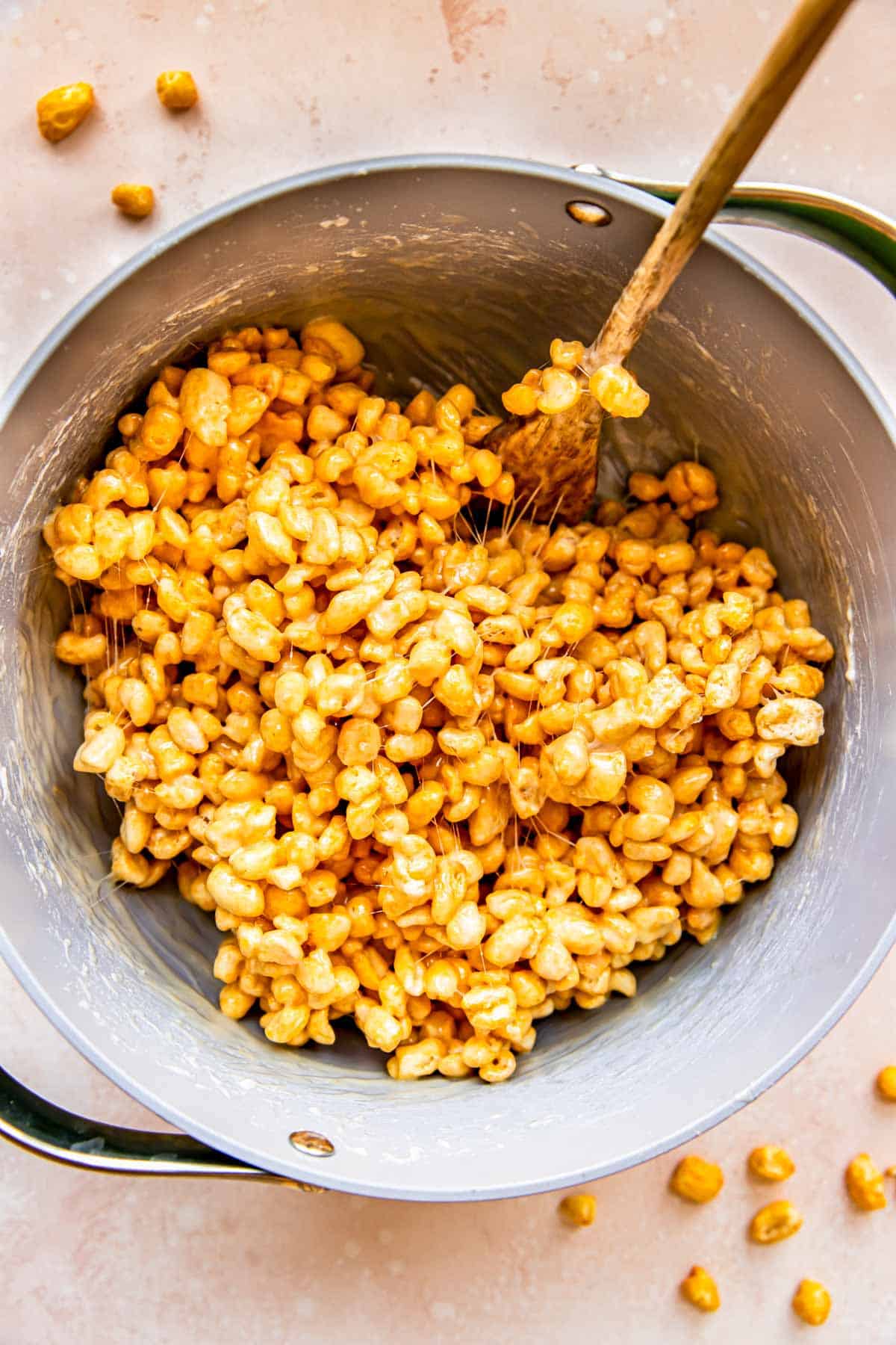 A wooden spoon is stirring cereal and the marshmallow mixture together. 