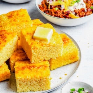 Slices of cornbread stacked on a plate with bowls of chili in the background.
