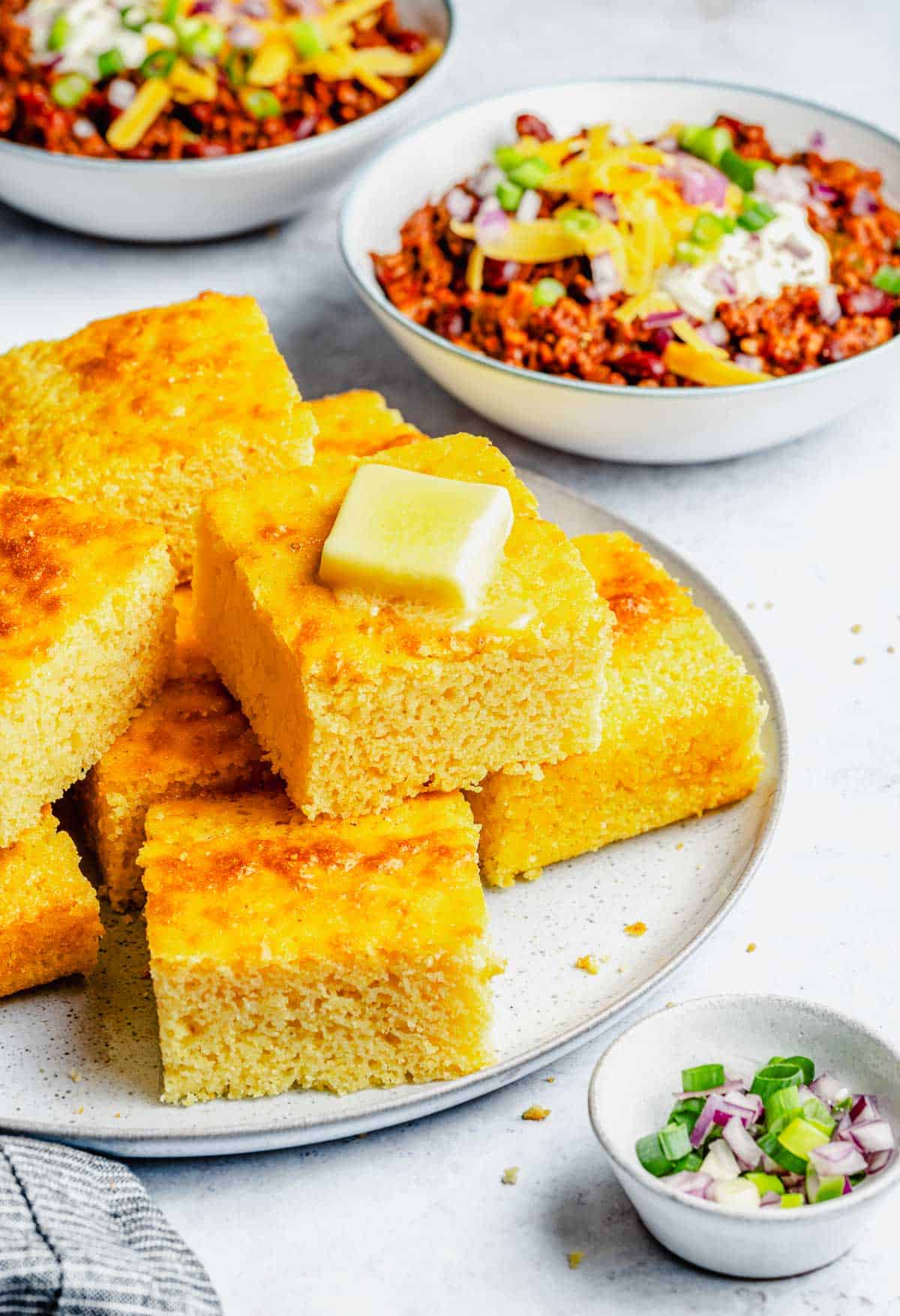 This cornbread recipe makes subtly sweet, light, fluffy, crumbly cornbread every time. Smother it with butter and serve it with chili. Slices of cornbread stacked on a plate with bowls of chili in the background.