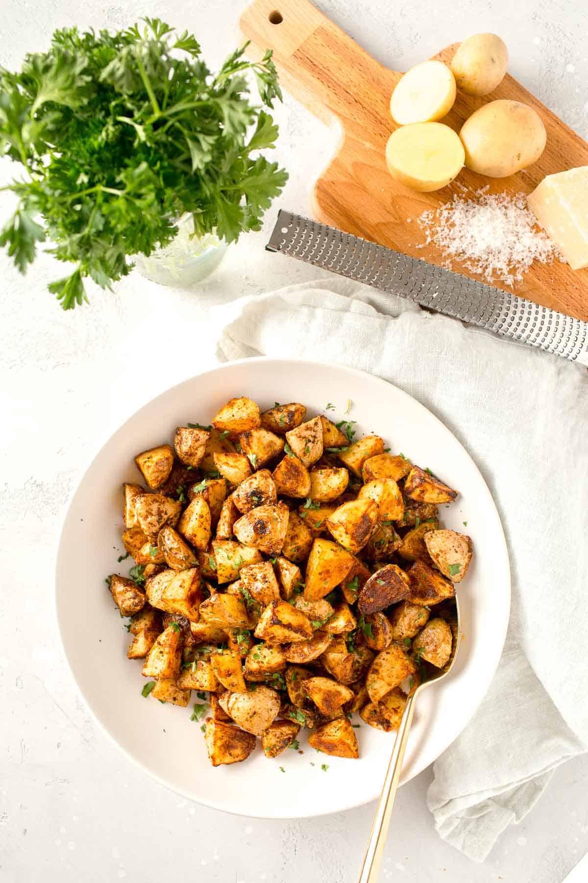 overhead image of roasted parmesan potatoes in a large white bowl next to a linen towel, fresh parsley, metal zester, and freshly grated parmesan cheese