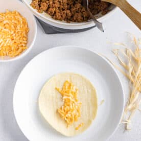 shredded cheese on a corn tortilla on top of a corn husk string on a white plate next to a bowl of ground beef