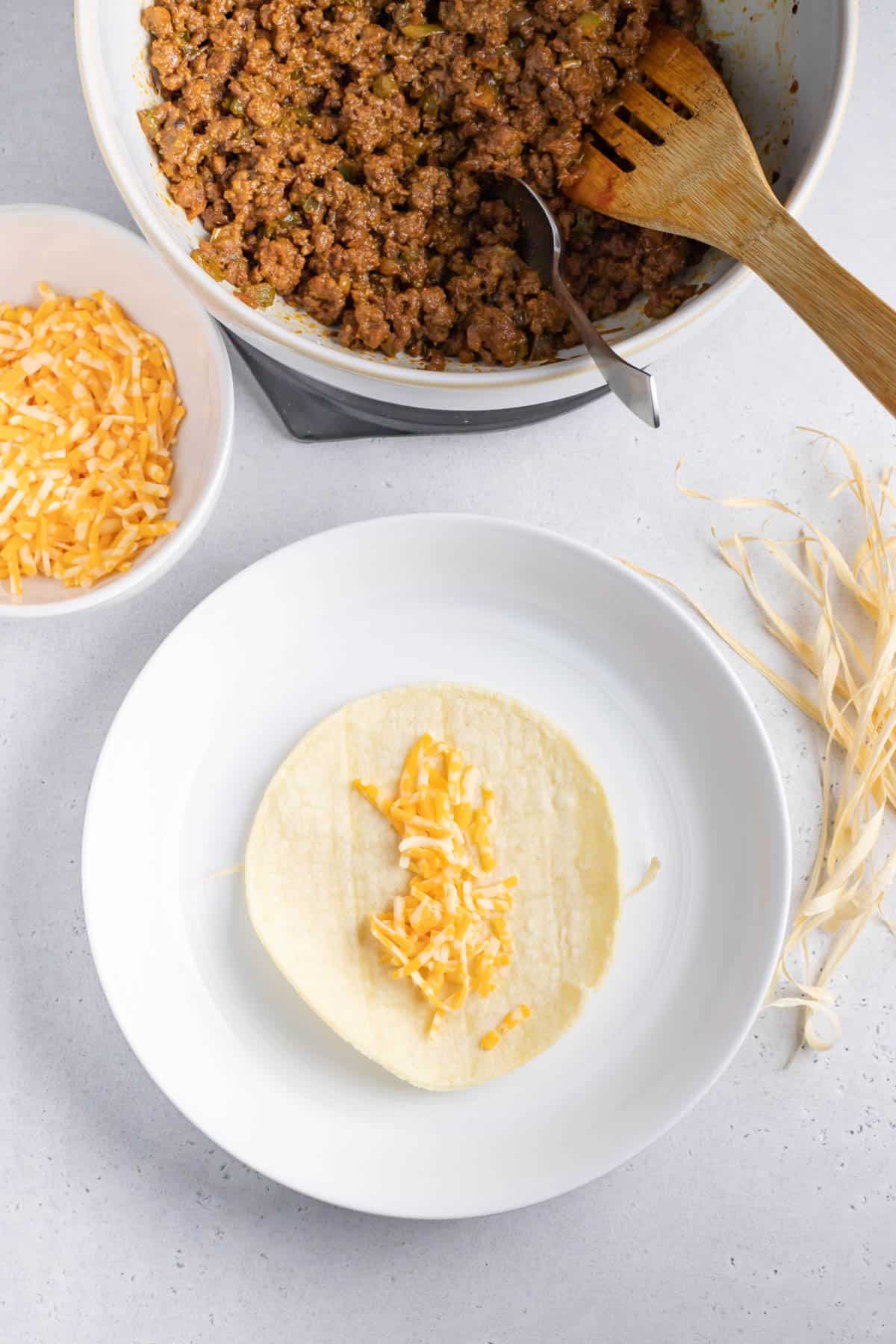 shredded cheese on a corn tortilla on top of a corn husk string on a white plate next to a bowl of ground beef