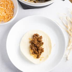 ground beef on top of shredded cheese on a corn tortilla on top of a corn husk string on a white plate next to a bowl of ground beef