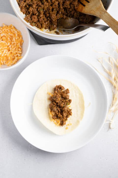 ground beef on top of shredded cheese on a corn tortilla on top of a corn husk string on a white plate next to a bowl of ground beef