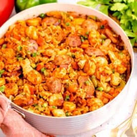 a large skillet of chicken and sausage jambalaya next to fresh ingredients and metal utensils