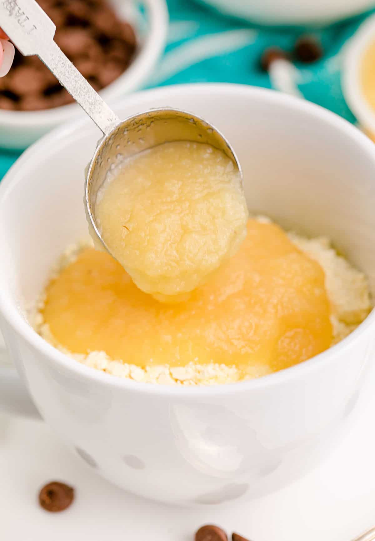 a tablespoon of apple sauce being added into a mug with cake mix