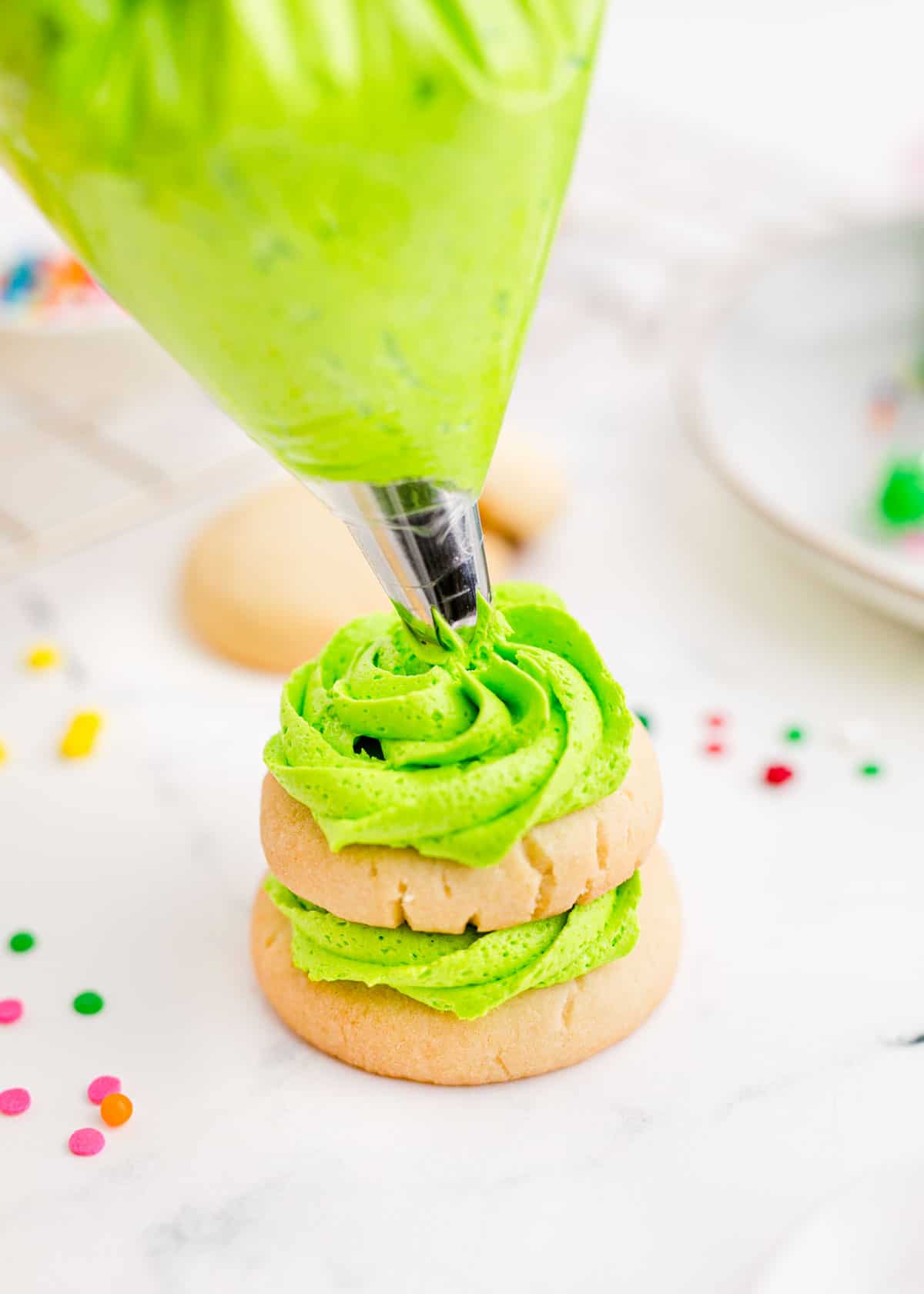Green icing is being piped onto a stack of sugar cookies to form a tree.
