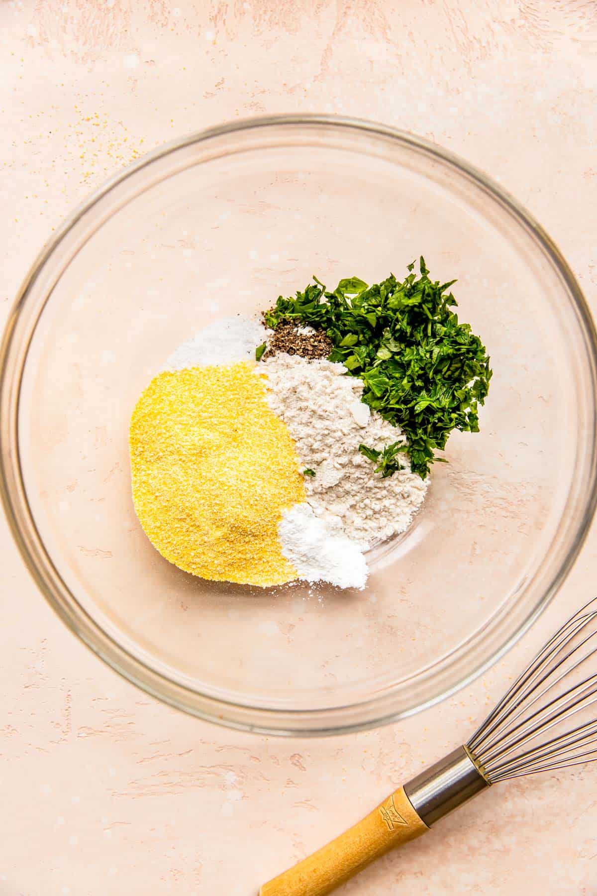 cornmeal, flour, fresh parsley, baking powder, salt, and pepper in a clear bowl next to a metal whisk
