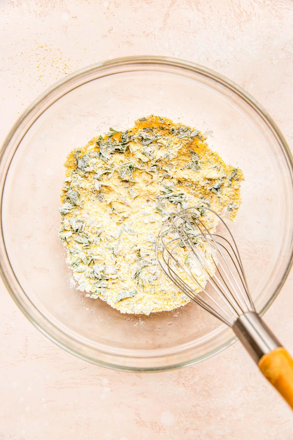 a metal whisk in a clear bowl mixing together cornmeal, flour, fresh parsley, baking powder, salt, and pepper.