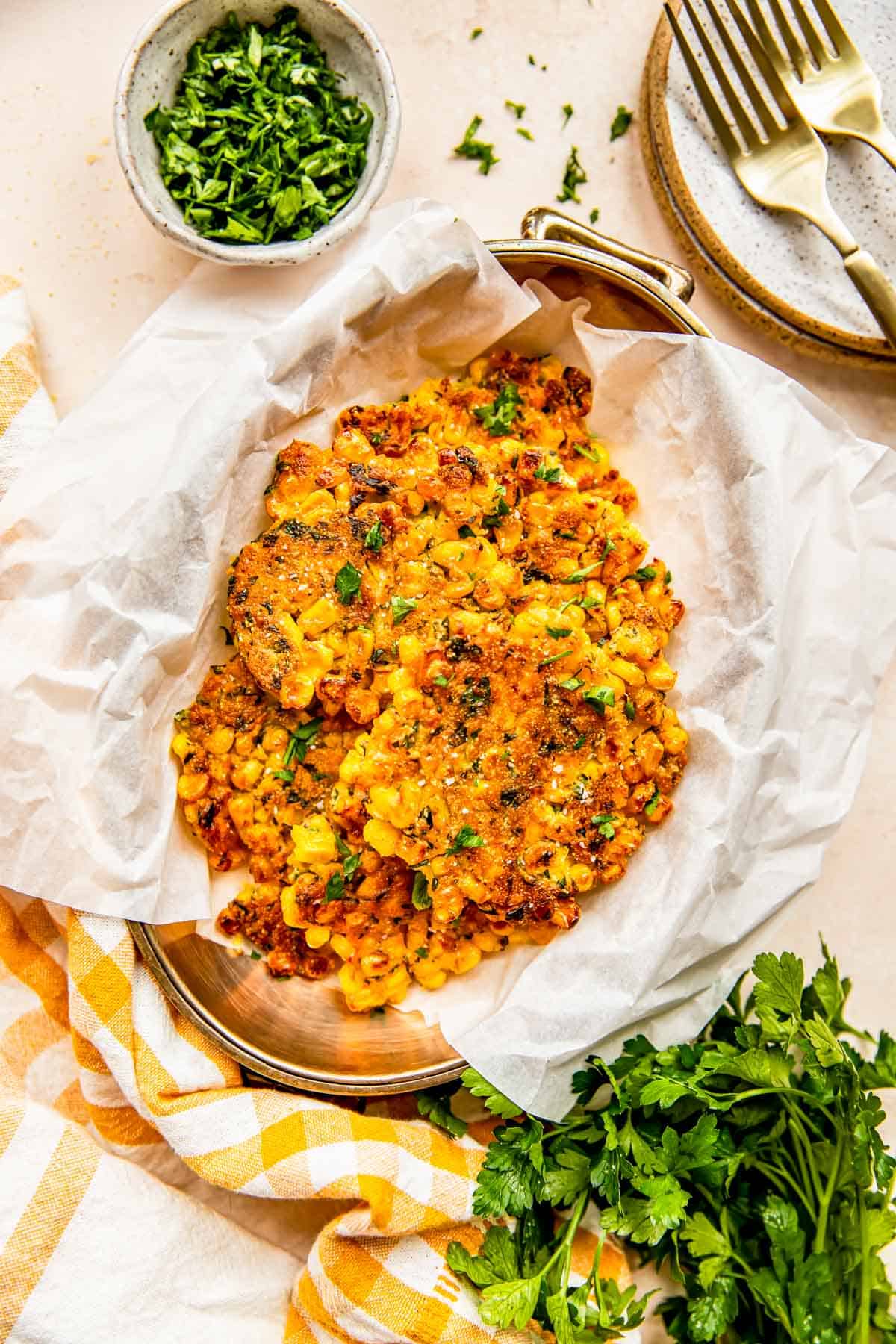 corn fritters on parchment paper in a metal container next to fresh parsley and a gingham linen towel