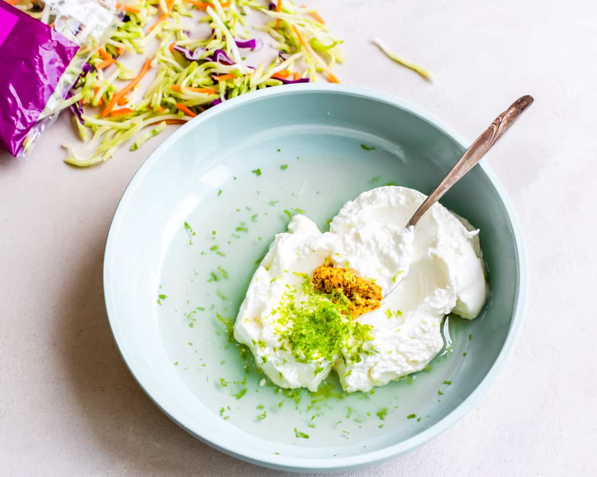 greek yogurt, lime zest, white vinegar, and dijon mustard in a light blue bowl with a metal spoon next to coleslaw mix