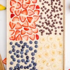 strawberry slices, chocolate chips, blueberries, and sliced bananas placed on top of pancake batter in metal sheet pan