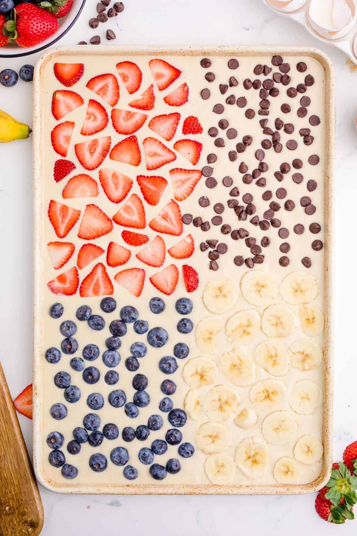 strawberry slices, chocolate chips, blueberries, and sliced bananas placed on top of pancake batter in metal sheet pan