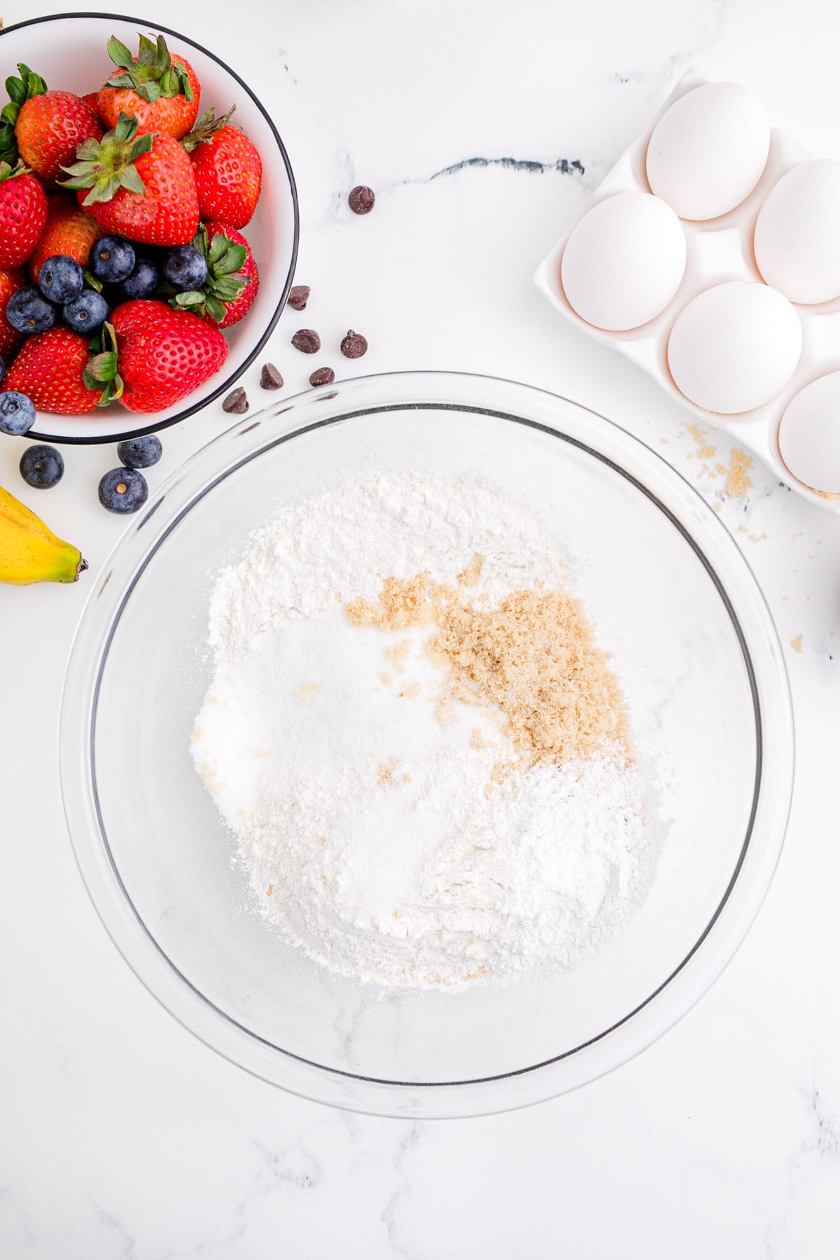 flour, white sugar, brown sugar, baking powder, and salt in a clear bowl next to fresh eggs and fresh berries