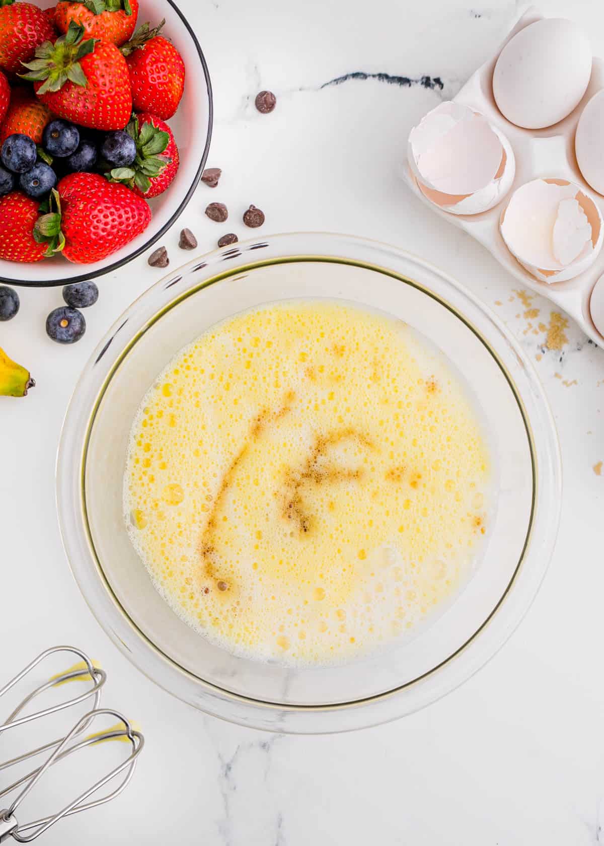 egg mixture with a splash of vanilla in a clear bowl next to broken egg shells and fresh berries