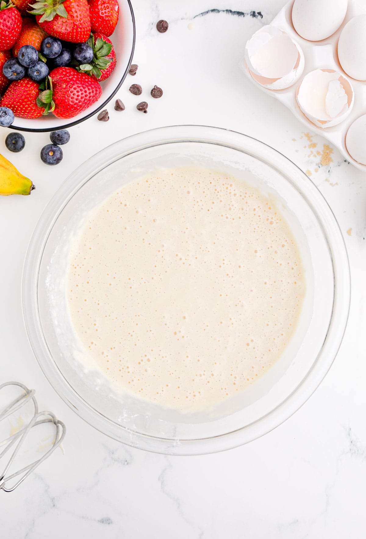 fruit pancake batter in a clear bowl next to broken egg shells and fresh berries