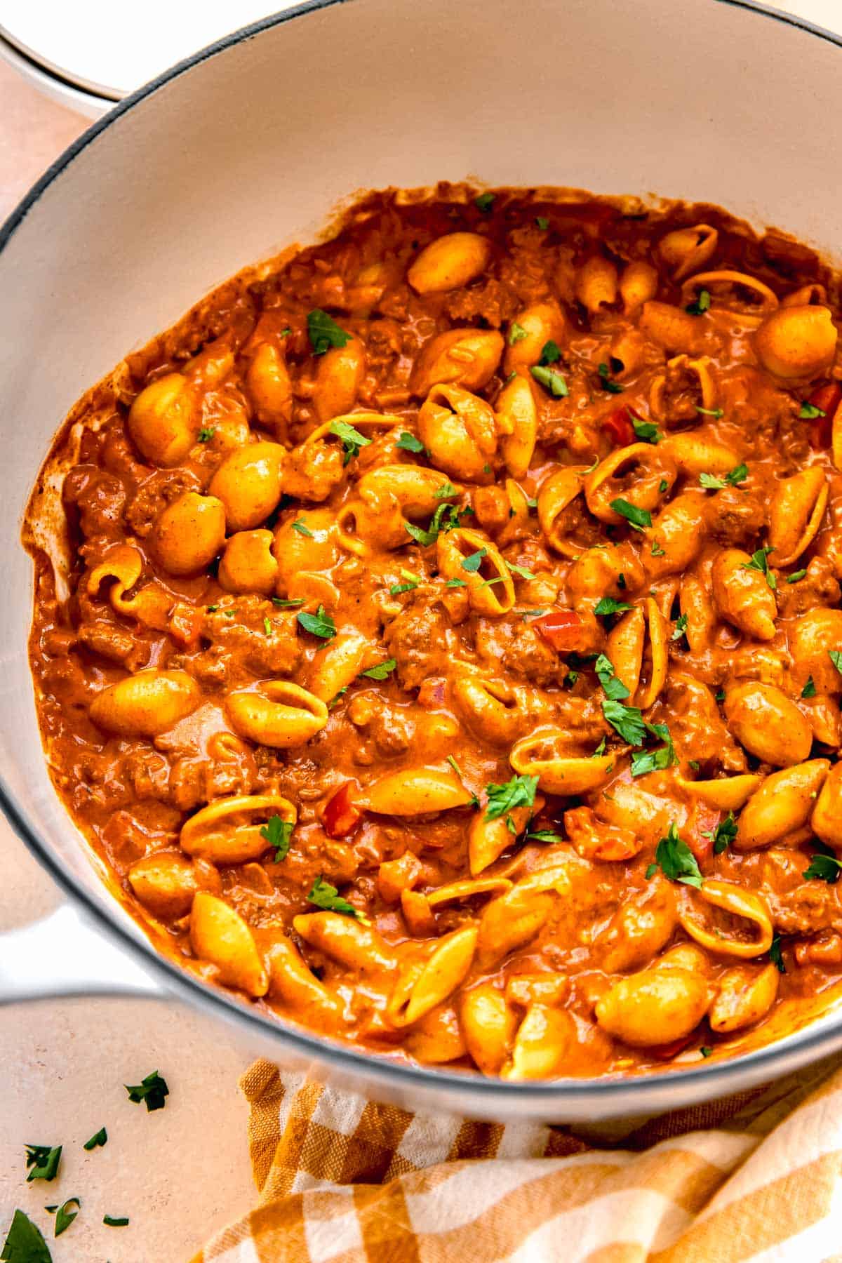 ground beef with pasta shells in a cast iron skillet next to a yellow checkered linen towel with fresh parsley garnished on top