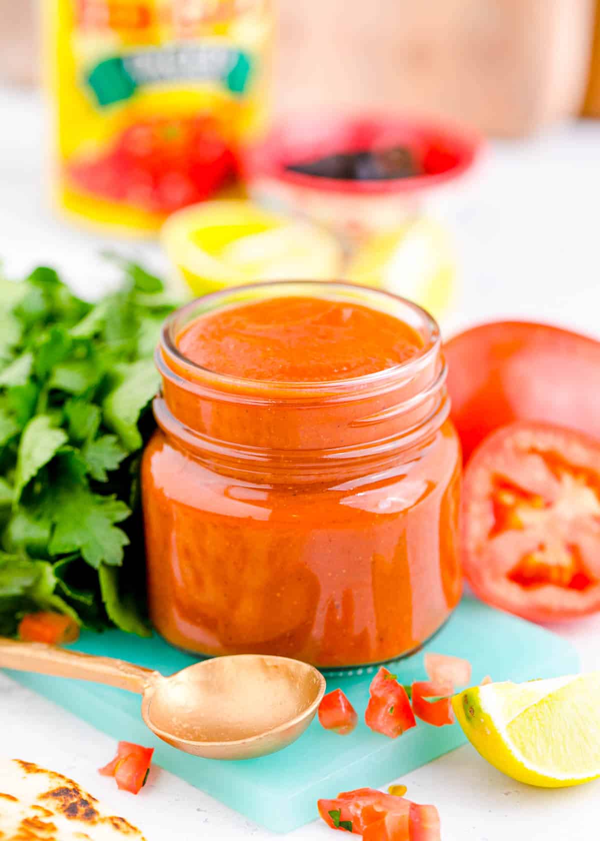 red enchilada sauce in a glass jar on top of a blue cutting board next to fresh ingredients