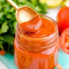 a spoon dips into red enchilada sauce in a glass jar on top of a blue cutting board