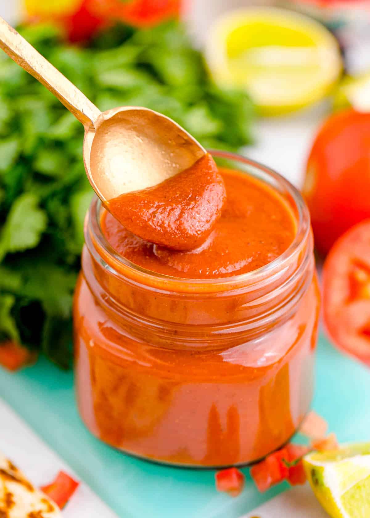 a spoon dips into red enchilada sauce in a glass jar on top of a blue cutting board