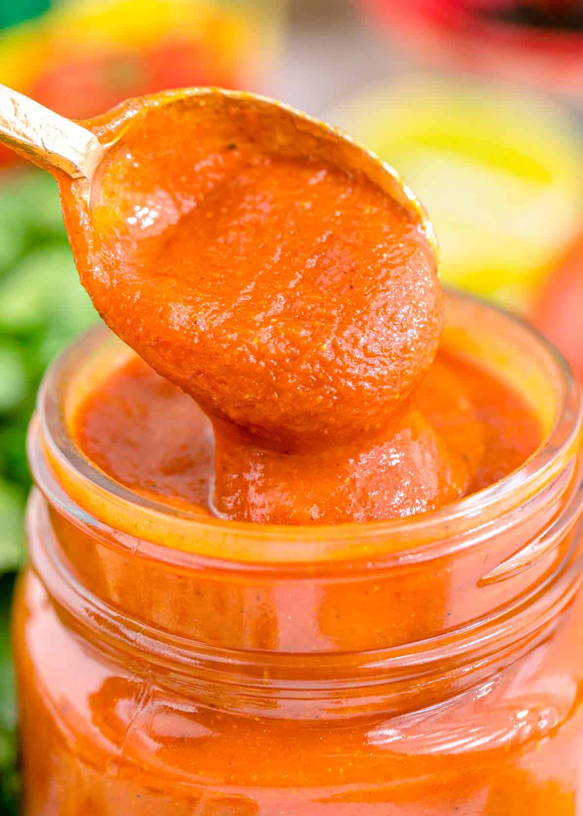 a spoon dips into red enchilada sauce in a glass jar