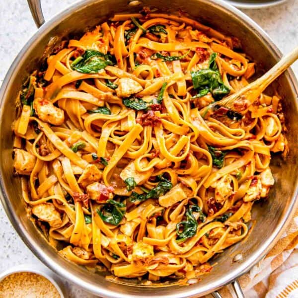 sun-dried tomato chicken pasta in a stainless steel skillet with a wooden spoon next to a yellow linen towel