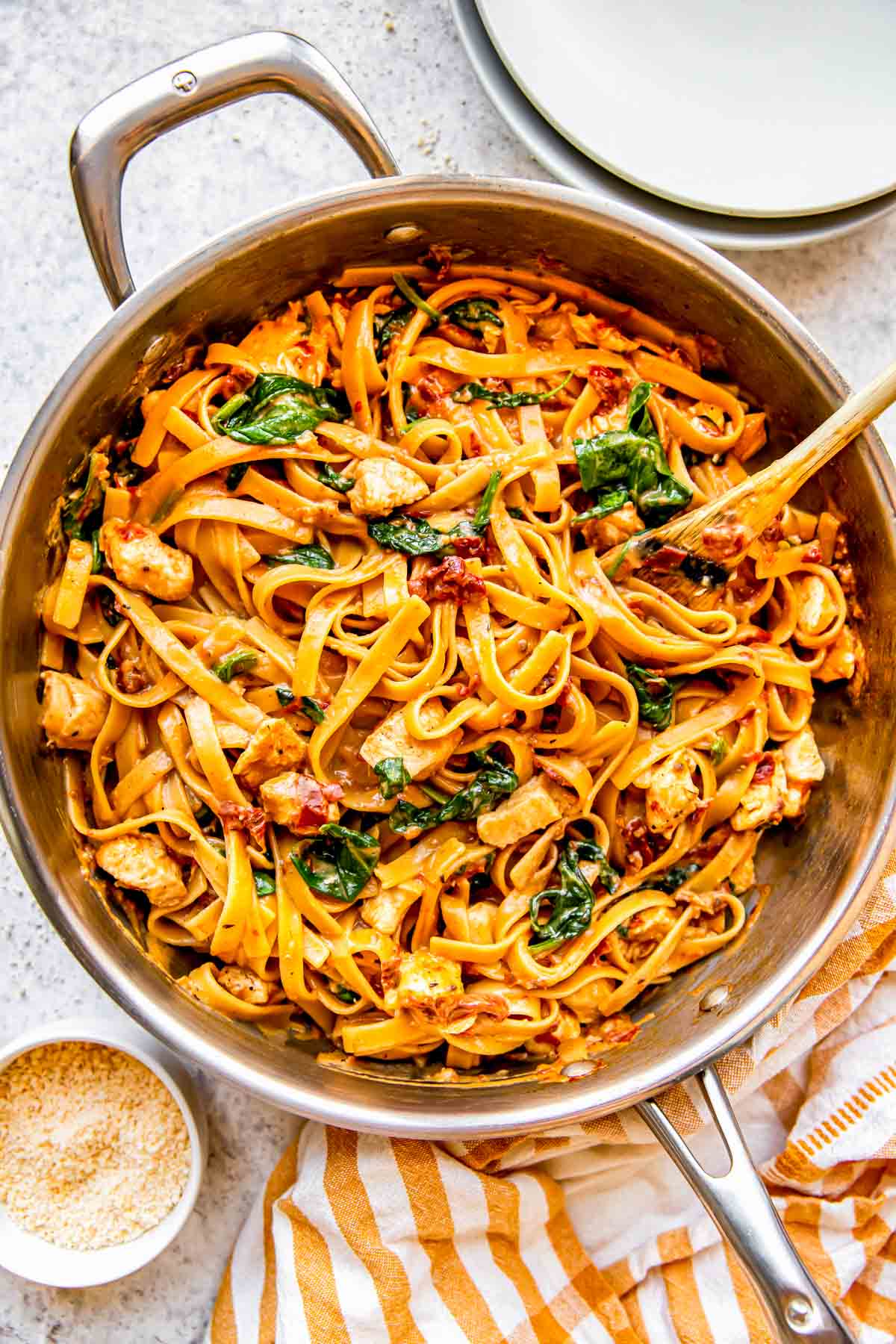 sun-dried tomato chicken pasta in a stainless steel skillet with a wooden spoon next to a yellow linen towel