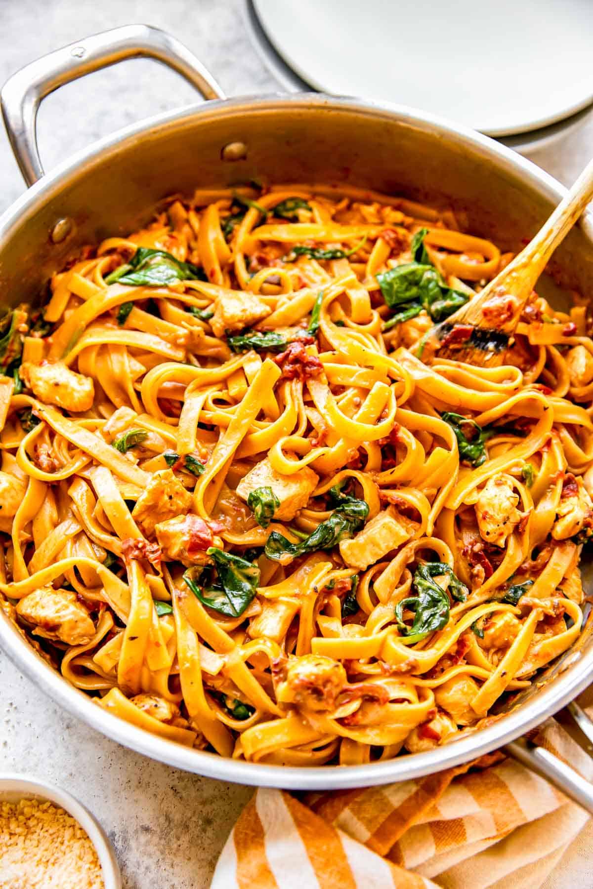 sun-dried tomato chicken pasta in a stainless steel skillet with wooden spoon next to a yellow striped linen towel