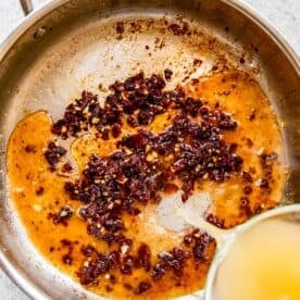 chicken broth being poured into stainless steel skillet with cooked garlic and sun-dried tomatoes