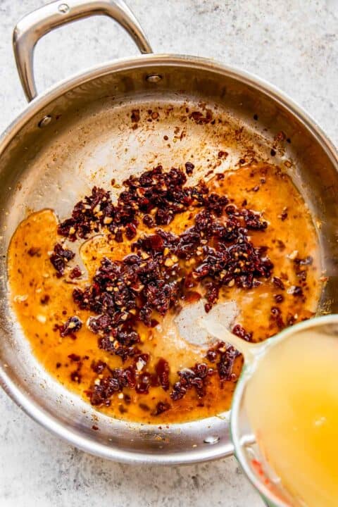 chicken broth being poured into stainless steel skillet with cooked garlic and sun-dried tomatoes