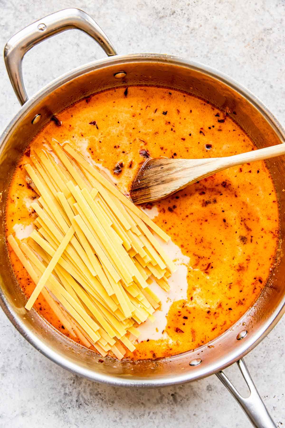 dry linguine pasta added to a stainless steel skillet with liquid and wooden spoon