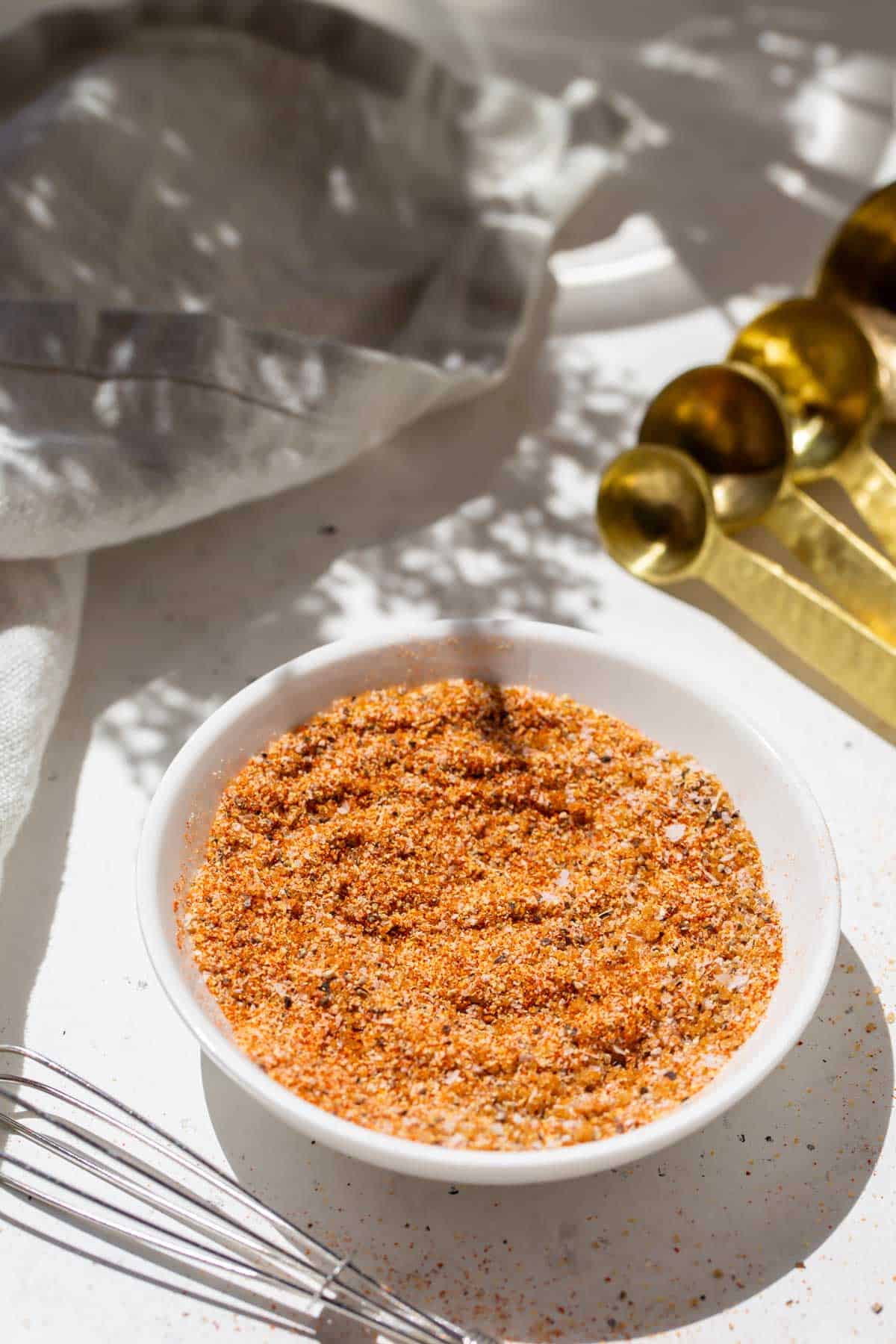 burger seasoning on a small white dish next to gold measuring spoons and a metal whisk