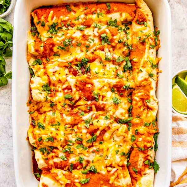 chicken enchiladas in a white baking dish with fresh cilantro garnished on top next to fresh ingredients like lime, sour cream, and red onions