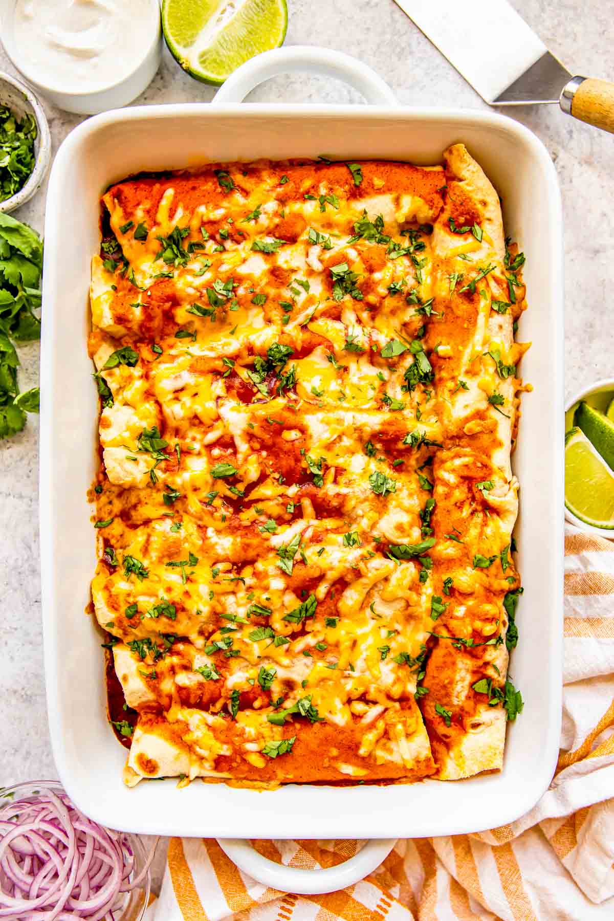 chicken enchiladas in a white baking dish with fresh cilantro garnished on top next to fresh ingredients like lime, sour cream, and red onions