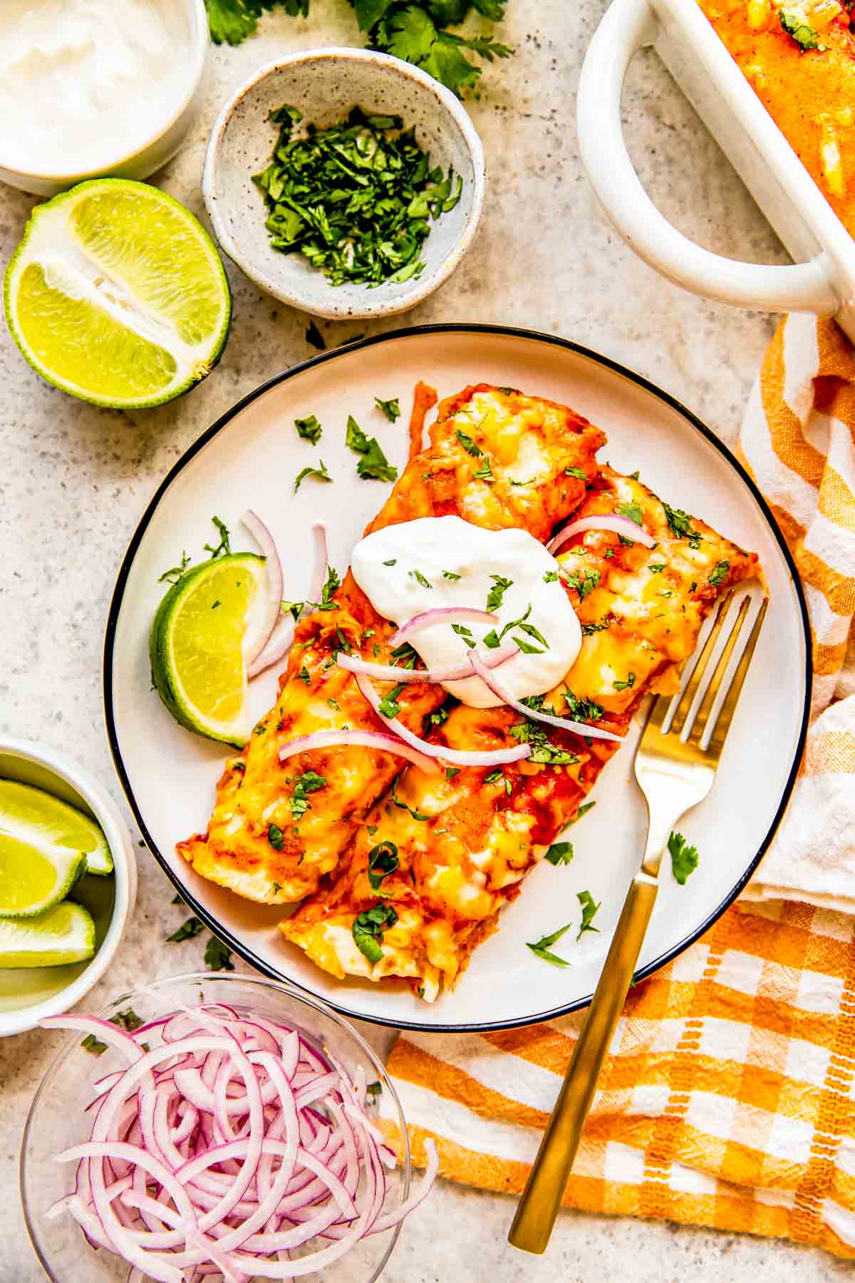 two chicken enchiladas on a plate topped with sour cream, red onions, and fresh cilantro. there is a lime and metal fork on the plate as well.