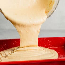 batter from a large clear bowl is being poured into a red baking dish
