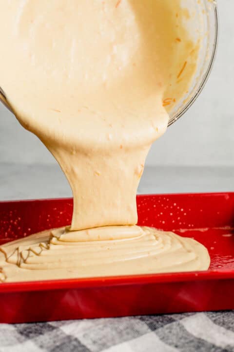 batter from a large clear bowl is being poured into a red baking dish