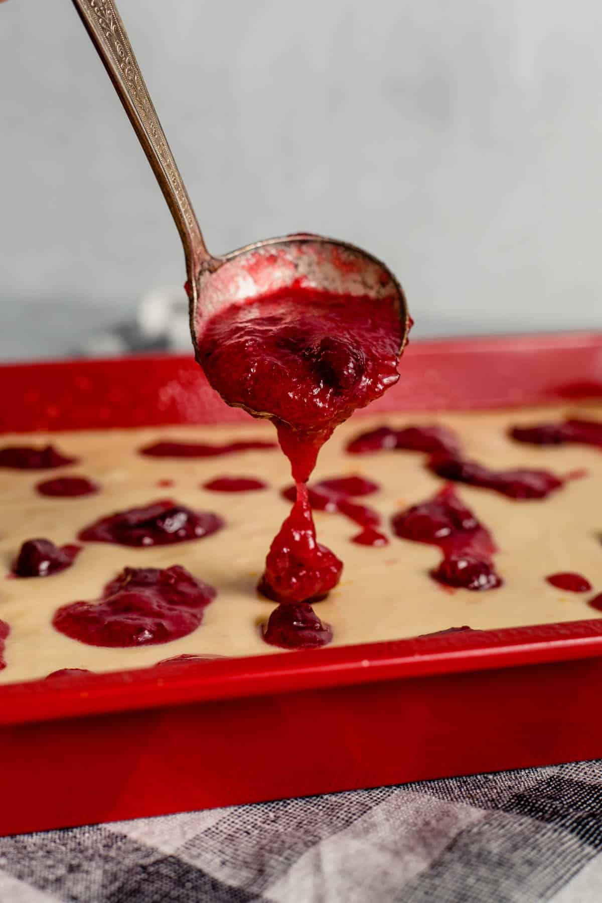 cranberry sauce is in a ladle being poured on top of the batter in the red baking dish