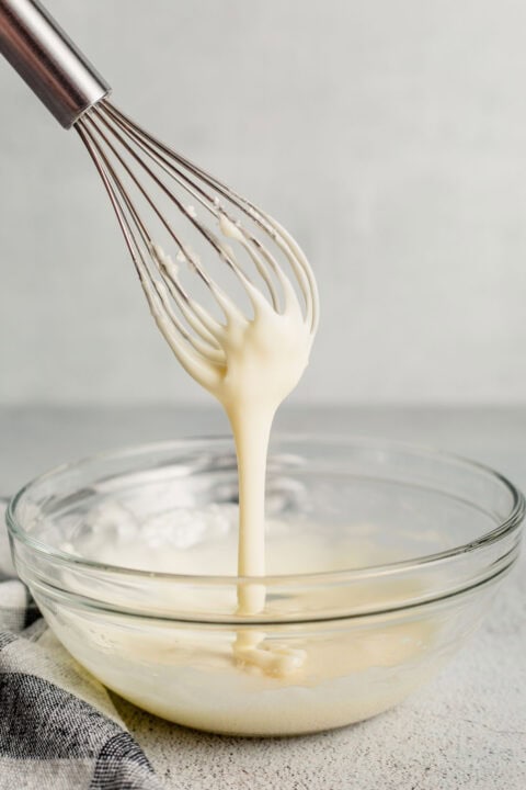 a whisk is being held above a small clear bowl with glaze running off the whisk