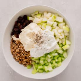 mayo and plain greek yogurt with freshly ground black pepper on top of diced apples, shallots, celery, pecans, cranberries, and shredded chicken in a large white bowl