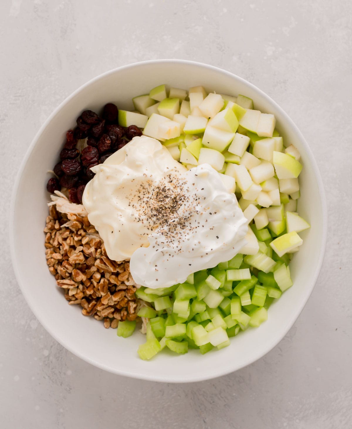 mayo and plain greek yogurt with freshly ground black pepper on top of diced apples, shallots, celery, pecans, cranberries, and shredded chicken in a large white bowl