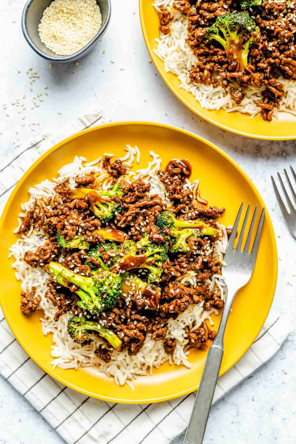 ground beef and broccoli plated on bright yellow plates on a bed of jasmine rice with a metal fork on the plate
