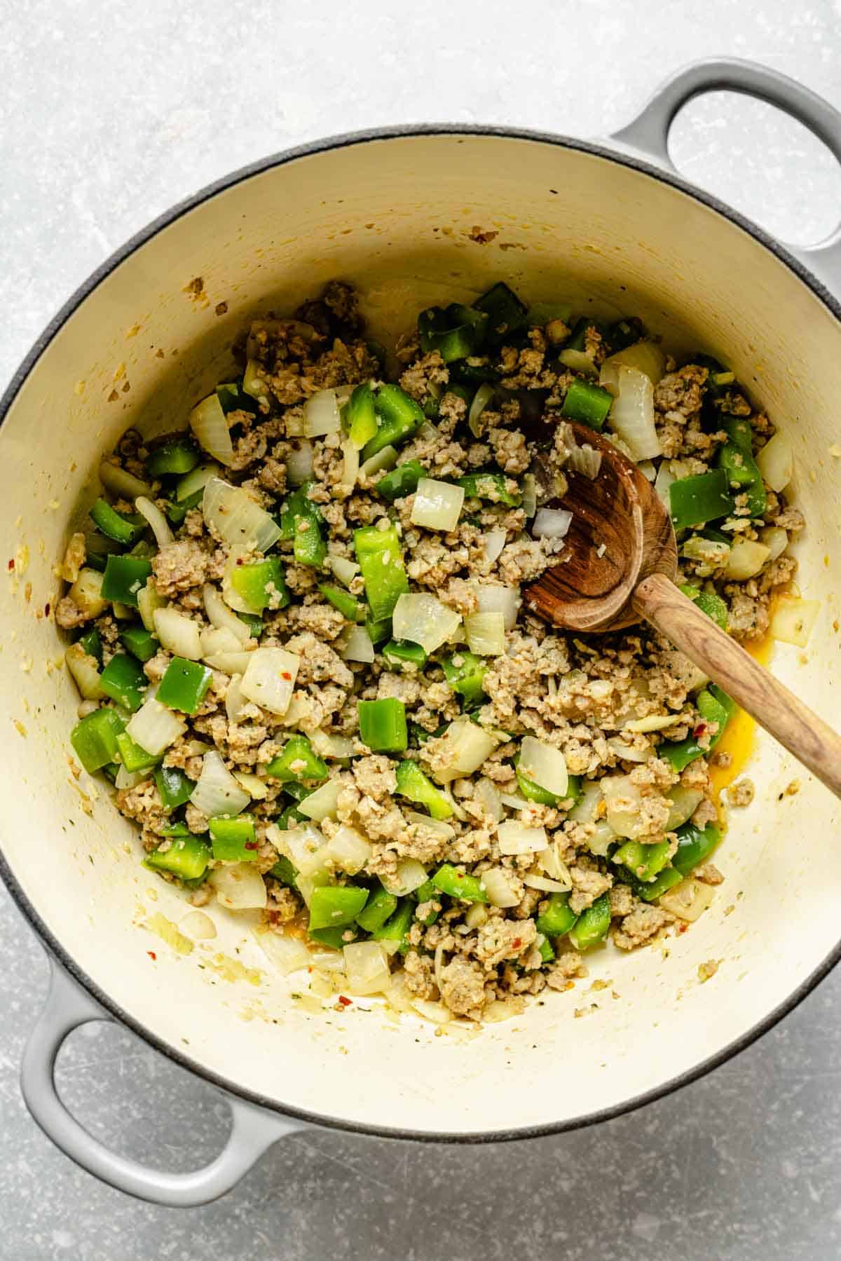 green bell peppers, onions, and crumbled sausage in a large dutch oven with a wooden spoon