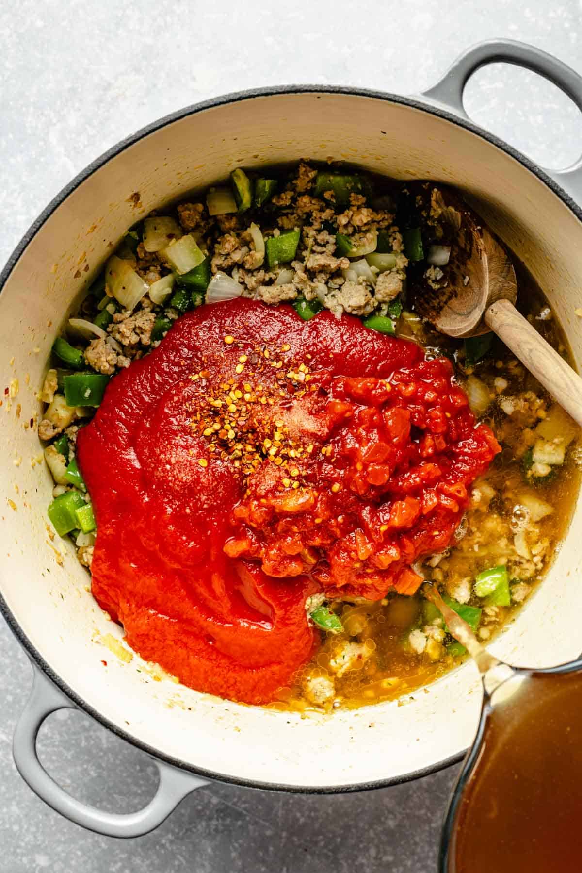 chicken stock being poured into the pot with tomato sauce, diced tomatoes, salt, and red pepper flakes added to sausage, onions, and bell peppers