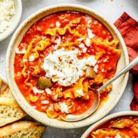 a bowl of lasagna soup with a dollop of ricotta cheese on top and a metal spoon next to toasted bread